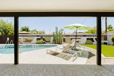 Covered patio framing a sunny Southwestern backyard with a curved swimming pool, white lounge chairs under a green umbrella, tall cactus, turf lawn, and a geometric mural on the boundary wall.