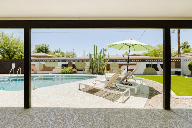 Covered patio framing a sunny Southwestern backyard with a curved swimming pool, white lounge chairs under a green umbrella, tall cactus, turf lawn, and a geometric mural on the boundary wall.