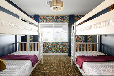 Cozy kids' bedroom with two white twin bunk beds facing a central window, colorful floral accent wall, navy curtains, patterned rug and burgundy throws on quilted mattresses.
