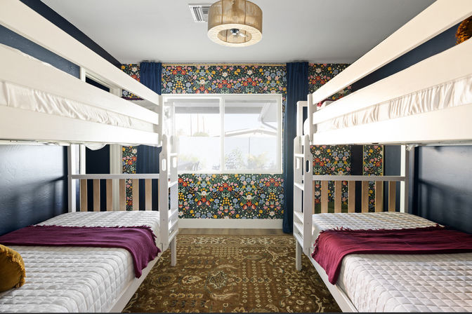 Cozy kids' bedroom with two white twin bunk beds facing a central window, colorful floral accent wall, navy curtains, patterned rug and burgundy throws on quilted mattresses.