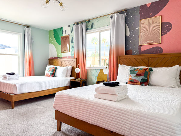 Bright modern guest room with two wooden platform beds, white quilts and folded towels, colorful geometric pillows, coral-ombre curtains, abstract mural accent wall and window with palm trees