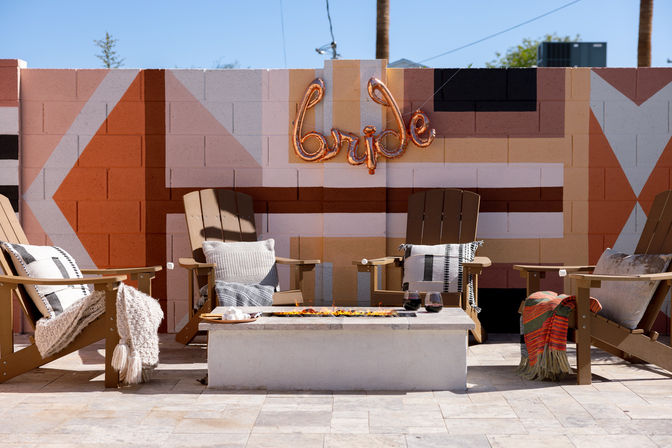Sunny outdoor patio with four wooden Adirondack chairs, cozy pillows and throws around a rectangular fire pit, colorful Southwestern geometric mural on a cinderblock wall and a rose-gold “bride” balloon sign.