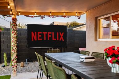 Backyard patio movie-night setup with long dining table, portable projector, black screen showing bold red streaming logo, string lights and floral-wrapped posts
