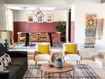 Open-concept living and dining room with terracotta half-painted accent wall, black dining table and wooden chairs, two mid-century armchairs with mustard pillows, round wood coffee table on plaid rug, woven wall hangings and potted plant — bright modern-boho interior.