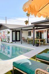 Sunlit mid-century modern backyard with turquoise pool, teal lounge cushions, covered dining patio, colorful floral mural, palm trees and orange umbrella.