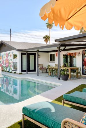 Sunlit mid-century modern backyard with turquoise pool, teal lounge cushions, covered dining patio, colorful floral mural, palm trees and orange umbrella.