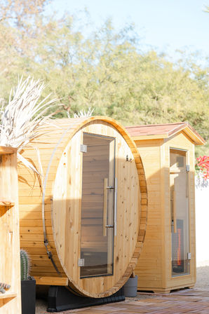 Outdoor wooden barrel sauna and adjacent cedar sauna cabin with glass doors on a sunny patio, desert-style landscaping with cactus and dry shrubs — cozy backyard spa scene.