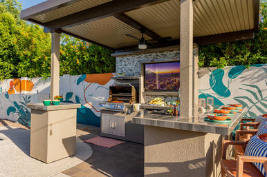 Modern covered outdoor kitchen and bar on a sunny backyard patio with a stainless-steel grill, wall-mounted TV showing a desert sunset, colorful mural, ceiling fan, and bar stools set with vibrant bowls.