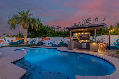 Resort-style suburban backyard pool at pink-purple sunset: curved blue pool with tiled steps, palm tree, colorful privacy-wall mural, covered outdoor kitchen with bar stools and lounge chairs.