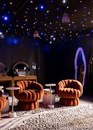 Moody boutique lounge interior with starry blue-and-white LED ceiling, two rust-orange plush shell chairs, marble-top gold side tables, animal-print rug, vanity mirrors and a purple backlit oval mirror.