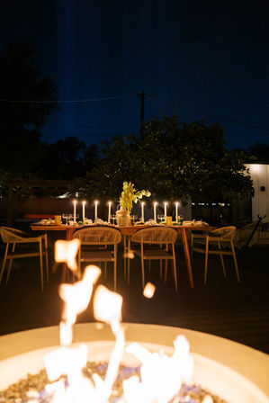 Cozy candlelit outdoor dining scene on a backyard patio at night — wooden table set with tall candles, woven chairs, a floral centerpiece, and a glowing fire pit in the foreground.