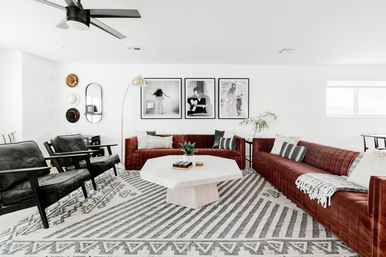 Bright contemporary living room with two tufted rust-red sofas and black leather armchairs arranged around a geometric white coffee table on a patterned gray rug, accented by a gold arc floor lamp, ceiling fan, and three black-and-white framed photos on a white wall.