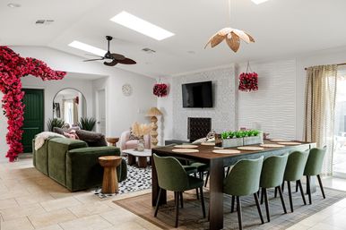 Sunlit open-plan living and dining room with vaulted skylights, green velvet sofa, wooden dining table with six green chairs, textured fireplace with wall-mounted TV, pink flowering arch and hanging baskets, rattan pendant lights and tiled floor