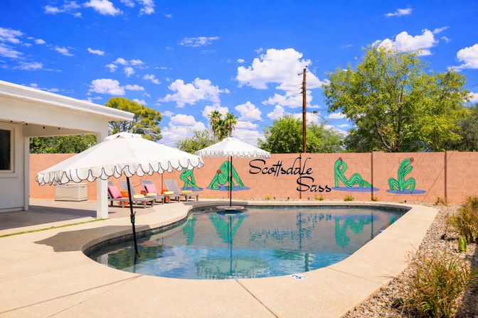 Sunny Scottsdale backyard pool with scalloped white umbrellas, colorful lounge chairs, pink cinderblock wall mural of stylized cacti, sparkling water reflecting a bright blue sky and desert landscaping.