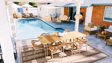 Sunny backyard patio oasis with a wooden outdoor dining table and wicker chairs under a pergola, rattan pendant lights, lounge seating and a blue in-ground swimming pool.