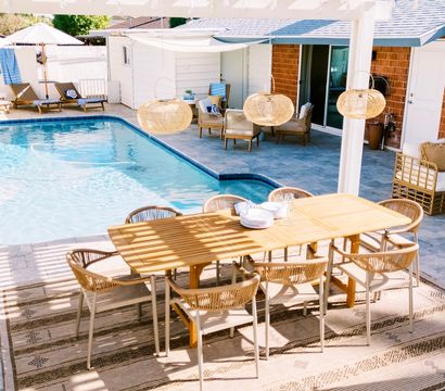 Sunny backyard patio oasis with a wooden outdoor dining table and wicker chairs under a pergola, rattan pendant lights, lounge seating and a blue in-ground swimming pool.