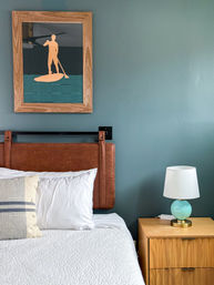 Cozy coastal-style bedroom with teal painted wall, leather headboard, crisp white bedding and striped throw pillow, framed paddleboard art above the bed and wooden nightstand topped with a turquoise lamp.