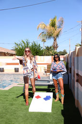 Two women in cowboy boots and sunglasses playing cornhole by a pool in a sunny backyard with palm trees