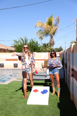 Two women in cowboy boots and sunglasses playing cornhole by a pool in a sunny backyard with palm trees