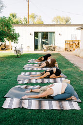 Four people doing child’s pose in an outdoor yoga class on a sunny backyard lawn, lined up on striped blankets and mats in front of a modern patio with sliding glass door.