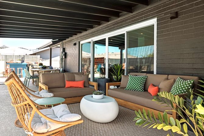Inviting covered backyard patio with wooden‑frame sofas and green patterned pillows, rattan lounge chairs, white oval coffee table, sliding glass doors and potted plants.