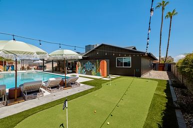 Resort-style backyard with turquoise pool, fringe umbrellas and wicker loungers, adjacent artificial-turf putting green, string lights, colorful mural on a dark house and tall palm trees under a clear blue sky.