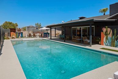 Sun-soaked rectangular backyard pool with turquoise water, covered patio with wicker seating and bar, umbrella-shaded lounge area, colorful cactus mural and palm tree under a clear blue sky