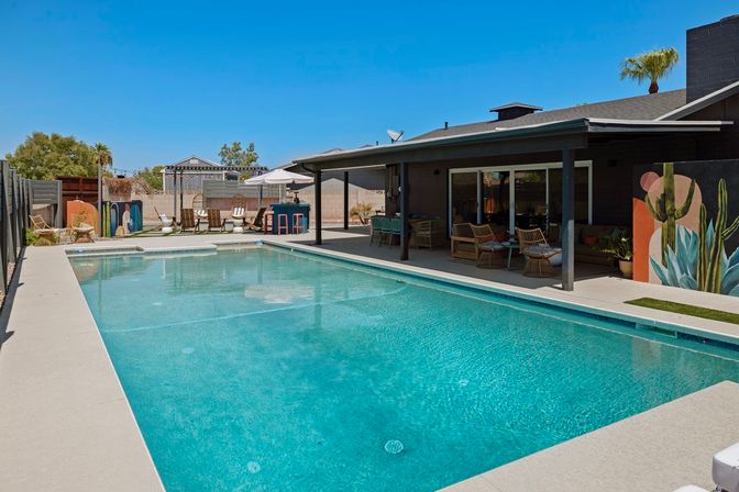 Sun-soaked rectangular backyard pool with turquoise water, covered patio with wicker seating and bar, umbrella-shaded lounge area, colorful cactus mural and palm tree under a clear blue sky