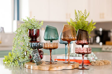 Colorful vintage stemmed glassware (amber, teal, ruby) arranged on a wooden board as a kitchen countertop centerpiece with leafy greenery and blurred cabinets in the background
