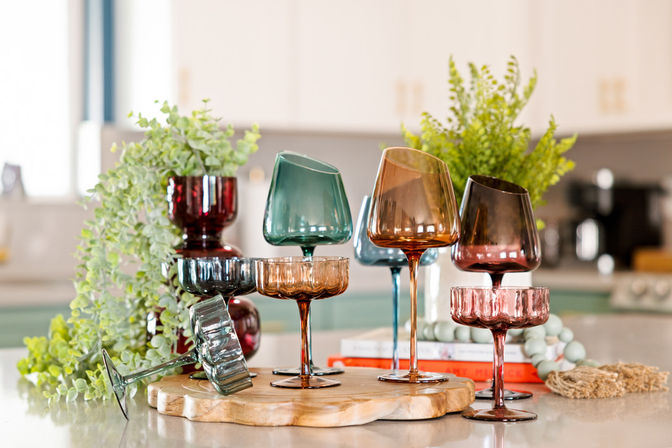 Colorful vintage stemmed glassware (amber, teal, ruby) arranged on a wooden board as a kitchen countertop centerpiece with leafy greenery and blurred cabinets in the background