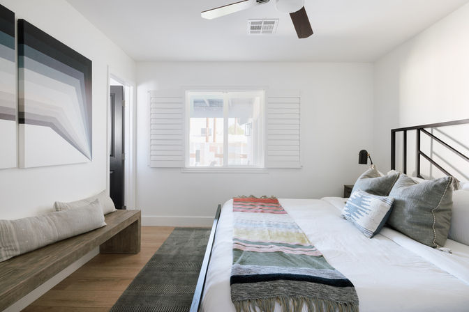 Sunlit minimalist bedroom with white walls and plantation shutters, king-size bed with patterned throw and gray pillows, black metal headboard, wooden bench, ceiling fan, and geometric wall art.