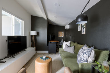Modern cozy living room with green sofa and patterned pillows, black accent wall, TV on white console, leather ottoman and a small home office nook under a sloped ceiling