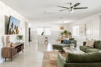 Sunlit modern open-plan living room and kitchen with coastal vibe: green velvet sectional, wooden coffee table and media console, woven poufs, wall-mounted TV showing palm trees, kitchen island with stools and dining area