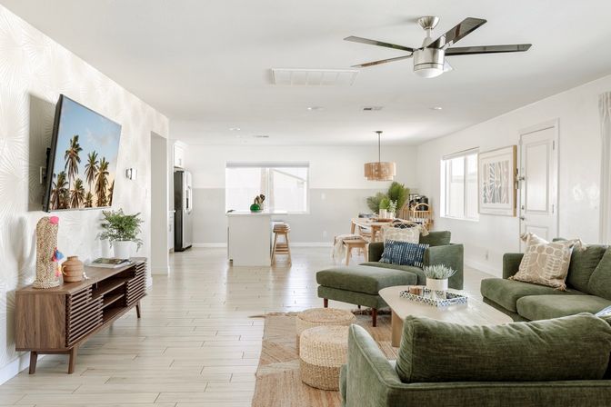 Sunlit modern open-plan living room and kitchen with coastal vibe: green velvet sectional, wooden coffee table and media console, woven poufs, wall-mounted TV showing palm trees, kitchen island with stools and dining area