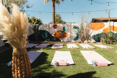 Sunny outdoor yoga setup on artificial turf with pink mats and blocks, abstract wall mural, palm tree and string lights