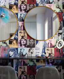 Bright beauty-studio vanity with pop-art tiled portraits of a woman, two gold-framed round mirrors, makeup brushes on a black counter, a ring light, and gray velvet chairs.
