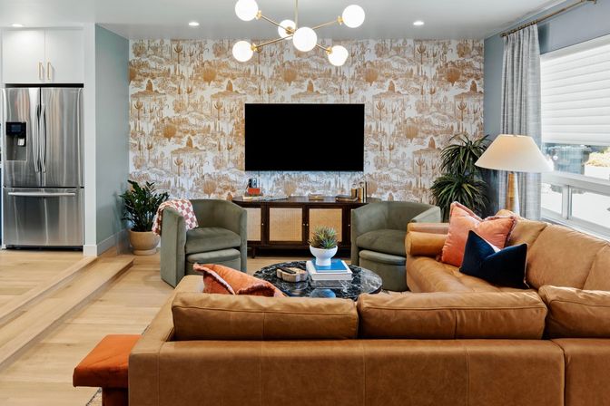 Open-concept living room with tan leather sectional, green lounge chairs, marble coffee table with potted plant, wall-mounted TV on patterned wallpaper, mid-century chandelier and stainless steel refrigerator visible in the adjacent kitchen.