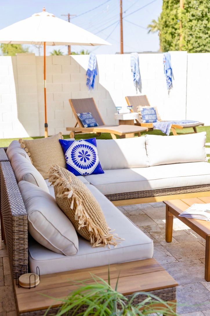 Sunlit backyard patio with wicker sectional sofa, neutral cushions and a blue patterned throw pillow, wooden coffee table, white umbrella and two chaise lounges on the lawn.