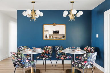 Eclectic dining nook with navy blue accent wall, gold-framed mirror reflecting a white kitchen, twin brass chandeliers, marble bistro tables and floral-upholstered chairs on light wood floor.