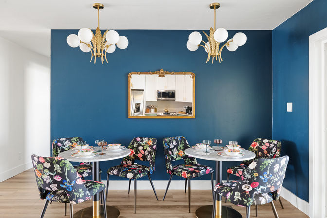 Eclectic dining nook with navy blue accent wall, gold-framed mirror reflecting a white kitchen, twin brass chandeliers, marble bistro tables and floral-upholstered chairs on light wood floor.