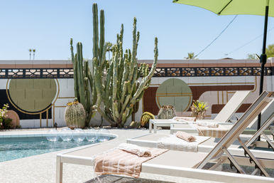 Sunny desert poolside with tall cacti, geometric mid-century mural, towel-draped lounge chairs and a green umbrella.
