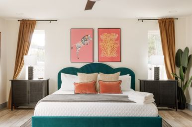 Bright modern bedroom with teal velvet scalloped headboard, white quilted bedding, coral accent pillows, pink zebra and abstract wall art, dark nightstands with lamps, gold curtains and a potted plant.