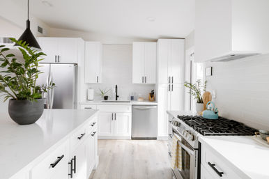 Bright modern white kitchen with large quartz island, black cabinet hardware, stainless steel refrigerator and dishwasher, gas range with teal kettle, pendant light and potted plants