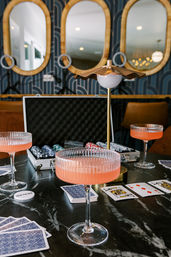 Ribbed coupe glasses with pink cocktails around playing cards and a poker chip case on a black marble table, lit by a gold table lamp with wicker-framed mirrors in the background — stylish indoor game night scene.