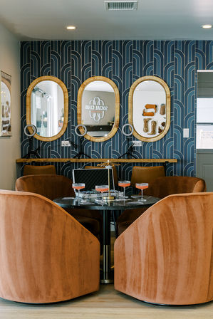 Cozy modern lounge with four terracotta velvet swivel chairs around a round marble table with cocktails, set against navy geometric wallpaper and three oval rattan-framed mirrors with ring lights.