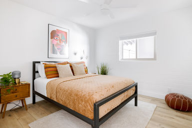 Bright modern minimalist bedroom interior with black metal bed frame, peach quilt and striped pillows, colorful abstract wall art, wooden nightstand with plant, window letting in natural light