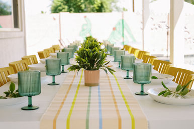 Sunlit outdoor dining table with yellow chairs, striped runner, potted green centerpieces, teal ribbed goblets and white plates topped with leafy sprigs.
