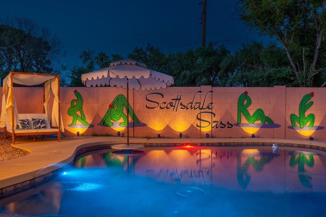 Nighttime Scottsdale backyard pool with colorful underwater lights, reflected scalloped umbrella, canopy daybed, and a lit wall mural of stylized green cacti and cursive lettering.