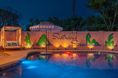 Nighttime Scottsdale backyard pool with colorful underwater lights, reflected scalloped umbrella, canopy daybed, and a lit wall mural of stylized green cacti and cursive lettering.
