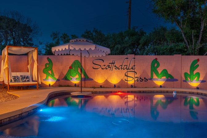 Nighttime Scottsdale backyard pool with colorful underwater lights, reflected scalloped umbrella, canopy daybed, and a lit wall mural of stylized green cacti and cursive lettering.